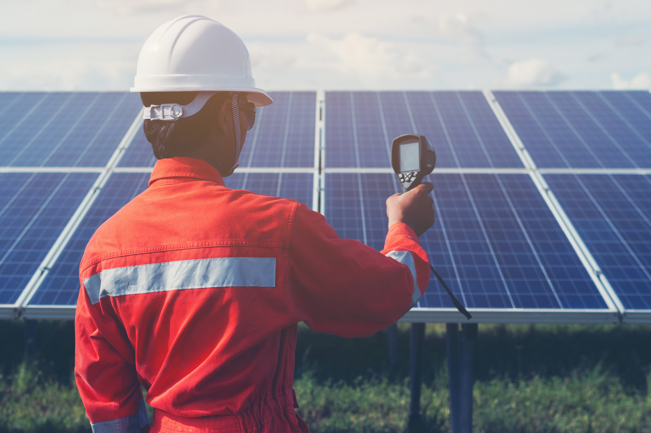 Man wearing hardhat and a red work jacket pointing a scanner at solar panels during the day